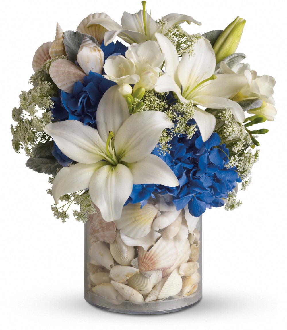 Everything's Beachy by Days Flowers Florida featuring blue hydrangea, white lilies, white freesia, Queen Anne’s lace, dusty miller, and a seashell beach scene inside an acrylic vase.