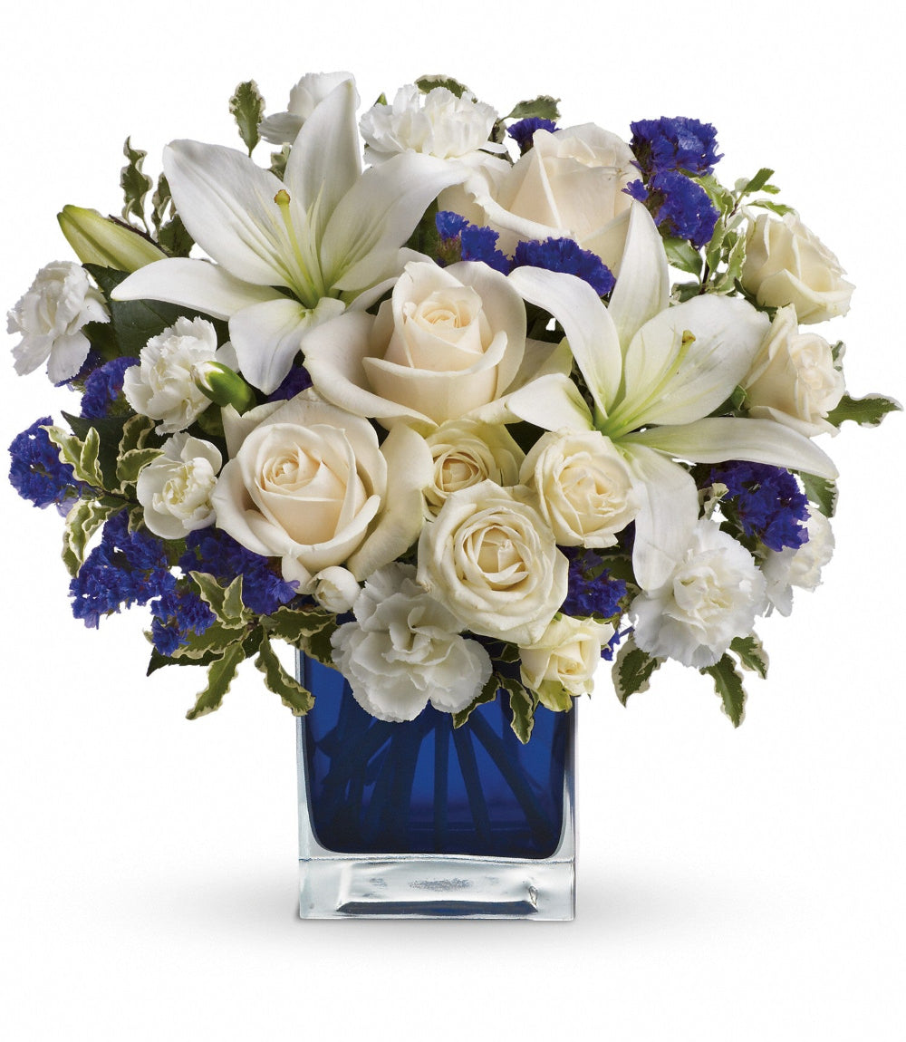 Sapphire Skies Bouquet by Days Flowers Florida featuring cream roses, white lilies, white miniature carnations, purple statice, and greenery arranged in a sapphire blue cube vase for an elegant and peaceful floral design.