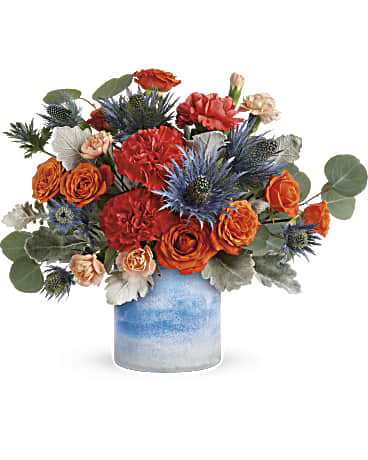 Standout Chic Bouquet by Days Flowers Florida featuring orange spray roses, dark orange carnations, peach miniature carnations, blue eryngium, seeded eucalyptus, and dusty miller in a frosted art glass cylinder vase.