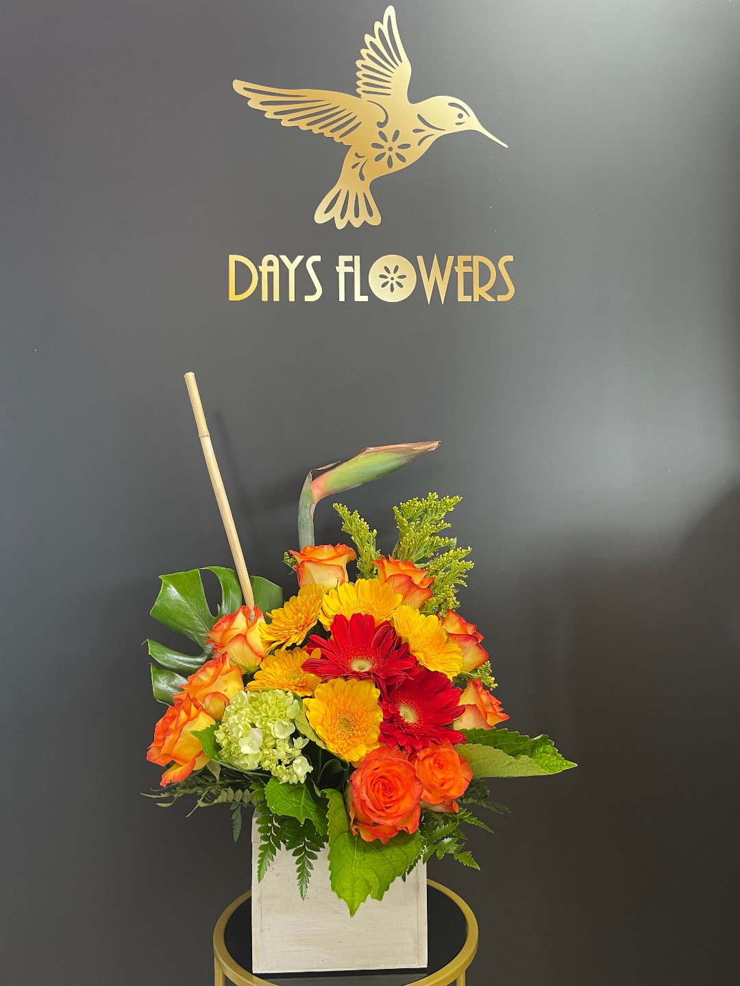 Vibrant floral arrangement by Days Flowers with red gerbera, orange roses, and green accents in a white square vase against a dark wall with gold logo.