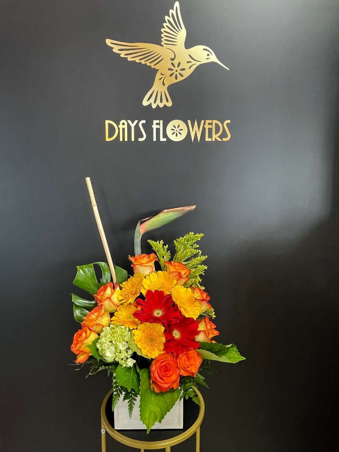 Elegant floral arrangement featuring bright oranges and reds with lush greens, showcased against a dark backdrop with ‘Days Flowers’ in gold lettering, perfect for Summerfield, Ocala, Orlando, The Villages, and Port St. Lucie deliveries.