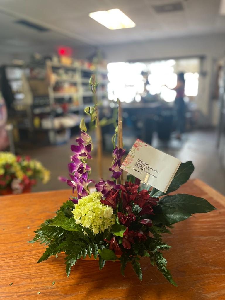 Elegant purple and dark red floral arrangement with lush greenery set against a blurred store interior, perfect for delivery in Summerfield, Ocala, Orlando, The Villages, and Port St. Lucie.