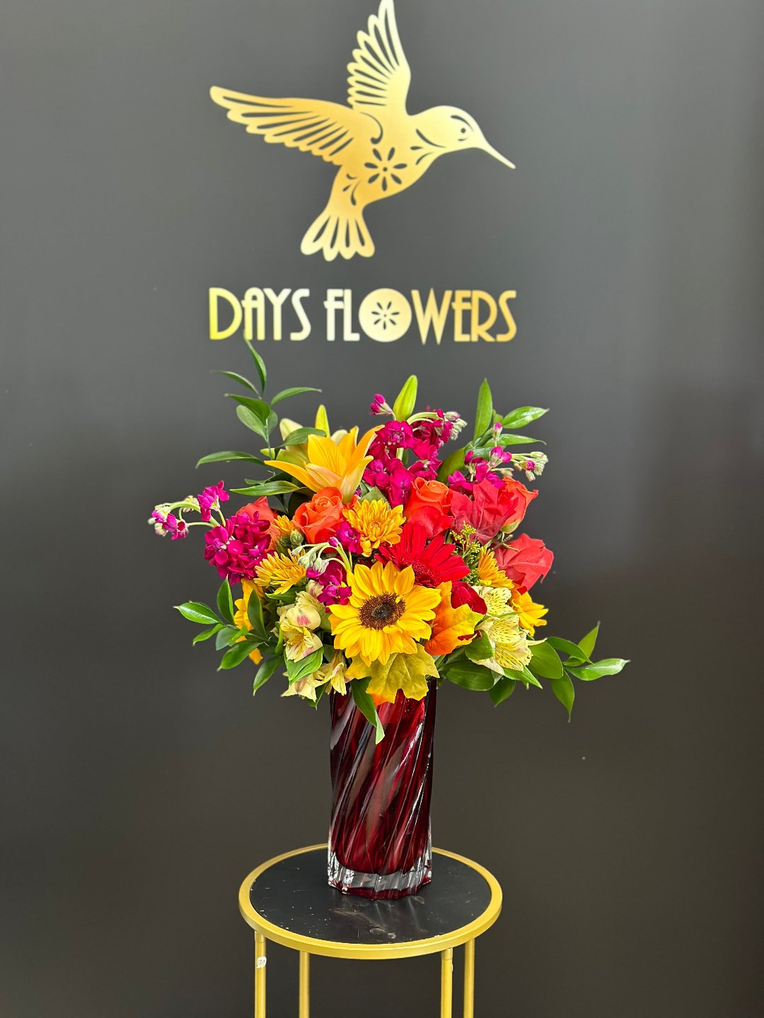 Close-up of the sleek red glass vase displaying fiery orange gerbera daisies and fresh botanical greenery