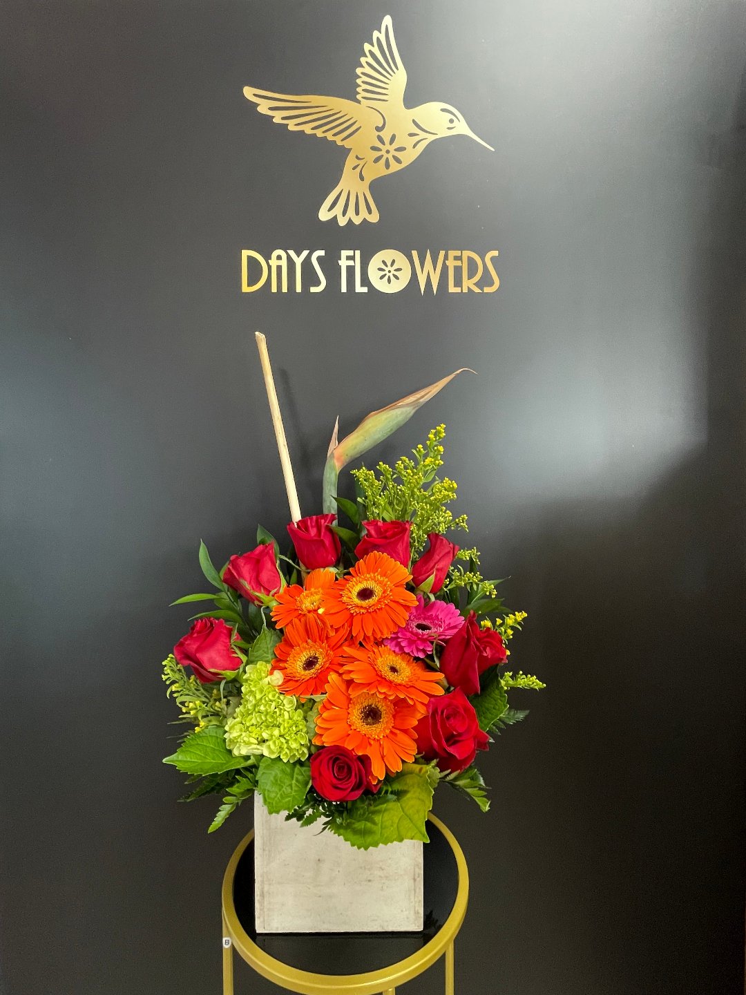 Elegant floral arrangement featuring vibrant red roses, bright orange gerberas, and lush greenery in a chic white vase against a dark backdrop with ‘DAYS FLOWERS’ in gold lettering.